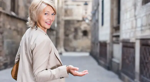 Uśmiechnięta kobieta z blond włosami w modnych, odmładzających krótkich fryzurach bob, zaprasza gestem ręki do wąskiej uliczki.