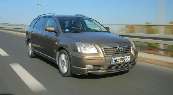 Brązowy Toyota Avensis T25 w kombi jedzie po drodze. Widać przednie żarówki i tablicę rejestracyjną WE 80036.