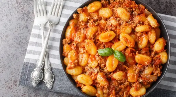 Un plat de gnocchi maison savoureux, nappés d'une sauce bolognaise riche et garnis d'une feuille de basilic frais. Deux fourchettes argentées sont posées sur une serviette à côté.