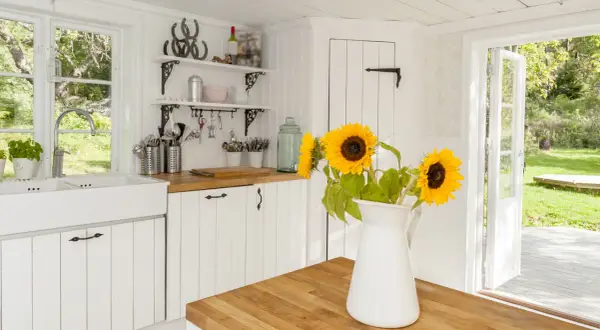 Helle Küche im modernen Landhausstil mit Spülbecken, Holzarbeitsplatte und Sonnenblumen. Offene Tür gibt Blick auf grünen Garten frei.