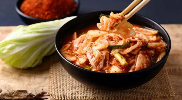 Miseczka pełna pikantnego kimchi, z pałeczkami podnoszącymi kawałek kapusty. Obok liść kapusty i miseczka z przyprawami. To właśnie kimchi!