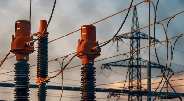Trzy izolatory z brązowymi głowicami, przygotowane do dystrybucji energii. W tle widać wieżę przesyłową, symbolizującą napięcie fazowe a międzyfazowe.