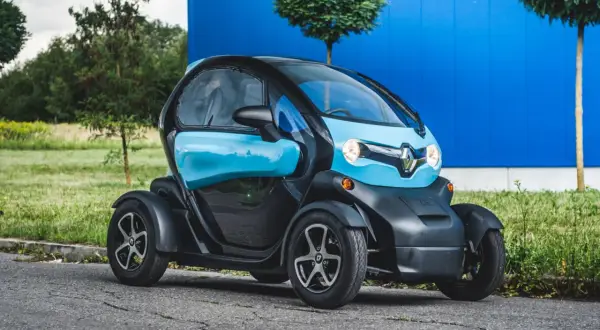 Niebiesko-czarny Renault Twizy, mały samochód elektryczny, zaparkowany na tle niebieskiego budynku i zieleni.