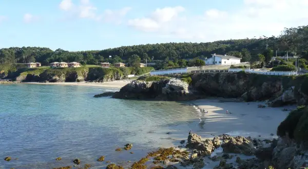 La **playa Burela** se asoma entre rocas y acantilados, con casitas blancas coronando la colina. El mar turquesa acaricia la arena dorada donde pasean algunas personas.
