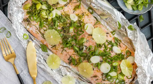 Lachsfilet in Alufolie gegrillt, garniert mit Limettenscheiben und Frühlingszwiebeln. Ein köstliches Gericht, das auf dem Grill zubereitet wird.