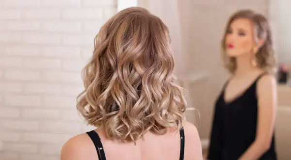 Kobieta z falowanymi włosami w odcieniu karmelowy blond, patrząca w lustro.