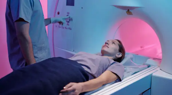 Une femme est allongée dans un appareil de TDM. Le technicien, en blouse bleue, ajuste les commandes. La TDM, c'est quoi ? Un examen d'imagerie médicale.