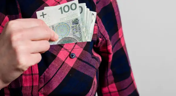 Ręka wkłada banknoty 100 zł do kieszeni koszuli w kratę. Może to symbolizować wynagrodzenie ryczałtowe.