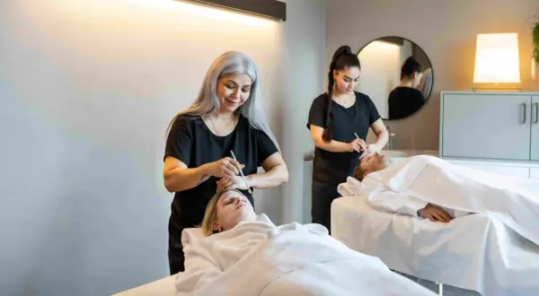 Dos terapeutas aplican tratamientos faciales a dos clientes en camillas. Una mujer con cabello plateado realiza un **olimpia masaje** facial a una cliente rubia.