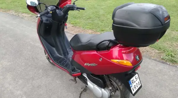Czerwony skuter Peugeot Elyseo 125 z czarnym kufrem na bagaż, gotowy na podróż po malowniczych drogach.