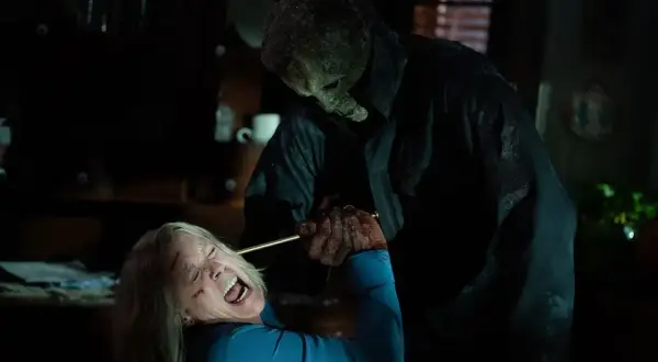 Michael Myers w masce i czarnym płaszczu trzyma nóż nad krzyczącą Laurie Strode. To przerażający halloweenowy finał.