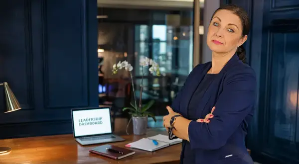 Kobieta w garniturze, z laptopem z napisem "LEADERSHIP DASHBOARD", uczy jak zostać managerem.