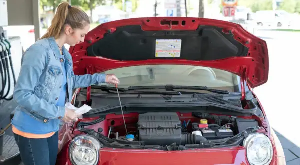 Junge Frau prüft mit einem Ölpeilstab den Motor eines roten Autos.