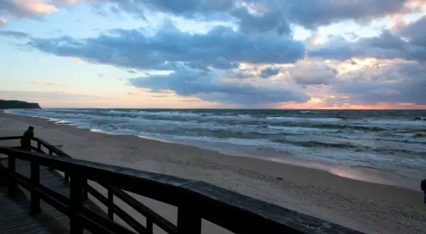 Zachód słońca nad morzem, plaża w Wiśle. Drewniany pomost prowadzi do piaszczystego brzegu, gdzie fale rozbijają się o brzeg.