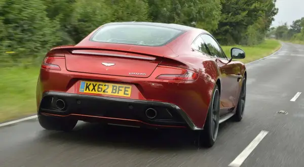 Czerwony Aston Martin Vanquish pędzi po mokrej drodze, otoczony zielenią.