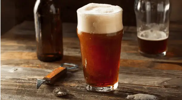 Un bicchiere di birra rossa con schiuma abbondante, accanto a una bottiglia e un apribottiglie. Perfetta per gustare i suoi gradi.