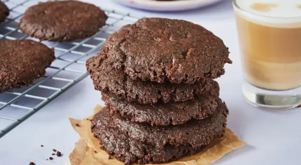 Ein Stapel saftiger brownie cookies, daneben ein Glas Latte Macchiato und ein Teller mit Schokoladenstücken.