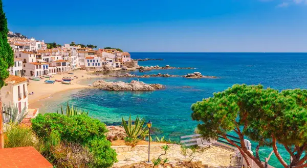 Vue pittoresque d'une crique ensoleillée avec des maisons blanches, des bateaux colorés et des eaux turquoises, parfait pour les activités Costa Brava.