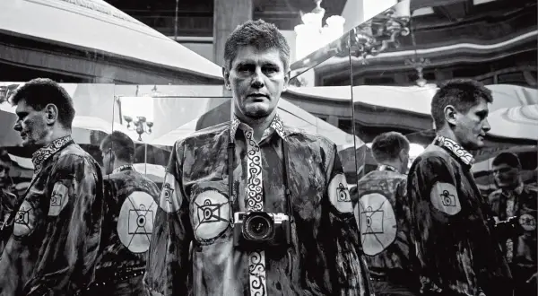 Krzysztof Miller w lustrzanym odbiciu, z aparatem na szyi, w koszuli z motywem oka.