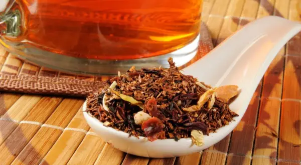 Suszone liście rooibos na łyżce, obok szklanki z bursztynowym naparem. Idealne do parzenia.