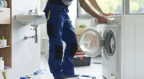 Instalator w niebieskich spodniach podłącza pralkę w łazience. Narzędzia na podłodze.