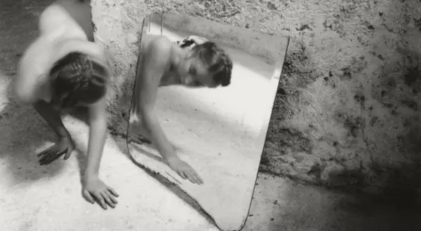 Une femme se contorsionne près d'un miroir brisé, son reflet fragmenté. Une œuvre de Francesca Woodman.