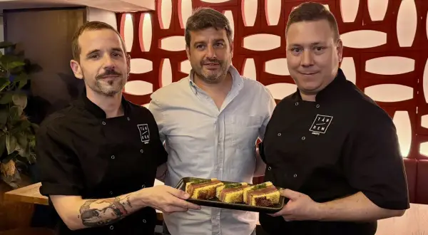 Tres hombres sonríen mientras uno sostiene una bandeja con aperitivos. ¡Un buen lugar donde comer en el Actur!