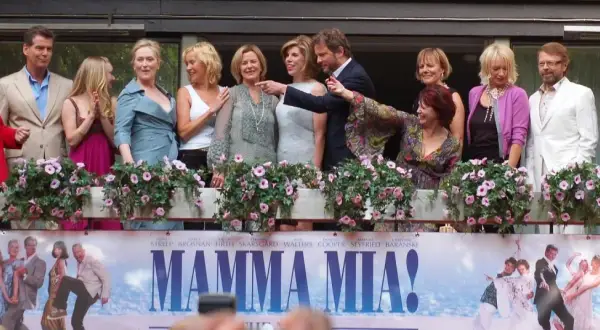 Obsada filmu "Mamma Mia!" pozdrawia fanów. W tle plakat z bohaterami, przypominający o wesołych piosenkach z filmu.