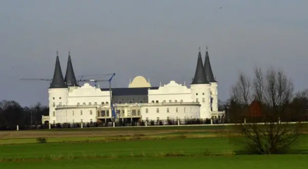Biały budynek przypominający zamek z ciemnymi wieżami, w tle żurawie budowlane.