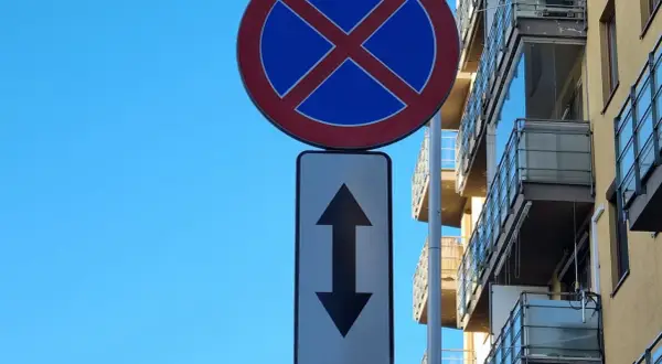 Znak zakaz postoju z tabliczką wskazującą, że zakaz obowiązuje na obu kierunkach. Budynek z balkonami w tle.
