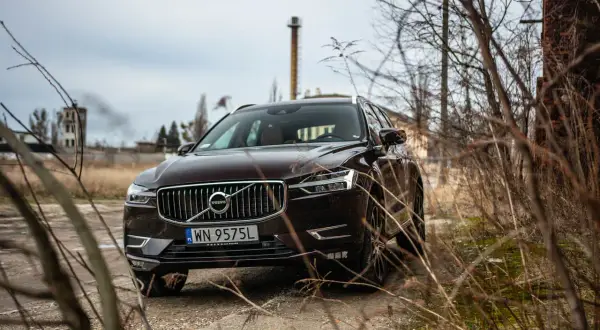 Brązowe Volvo XC60 T5 na tle opuszczonej fabryki i suchych gałęzi.