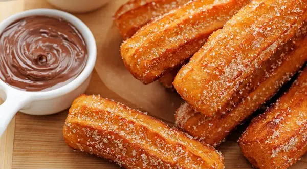 Chrupiące churrosy posypane cukrem, obok miseczka z gładką czekoladą. Idealna przekąska na słodko.