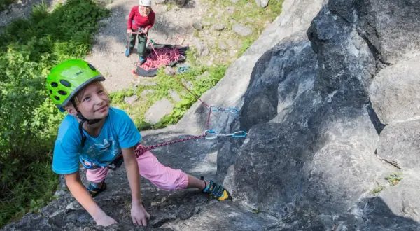 Ein Mädchen mit Helm klettert an einem Felsen, während eine andere Person sichert. Ein tolles Erlebnis beim **Klettern mit Kindern**.