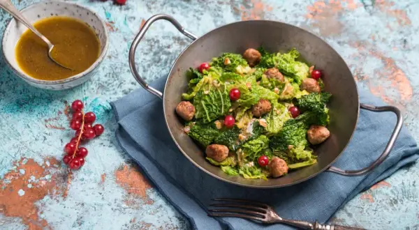 Wirsingsalat mit Fleischbällchen und roten Johannisbeeren in einer Metallschüssel, daneben eine Schüssel mit Dressing.