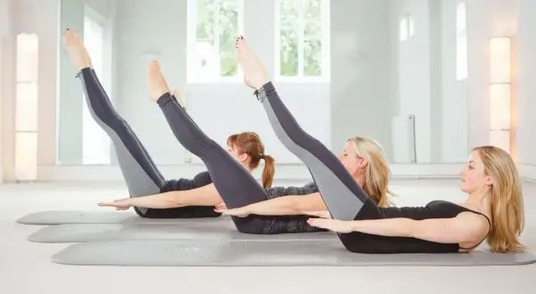 Tre donne eseguono posizioni pilates sul tappetino, sollevando gambe e braccia in un esercizio di core stability.