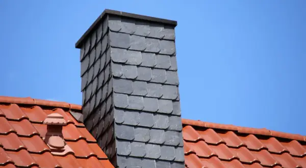 Moderner gemauerter Schornstein verkleidet mit grauen Schieferplatten, überragt ein rotes Ziegeldach unter blauem Himmel.