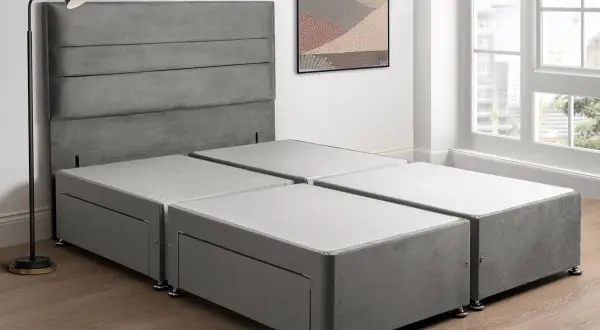 A grey sprung divan base with drawers, paired with a matching headboard, stands in a bright room with a large window.