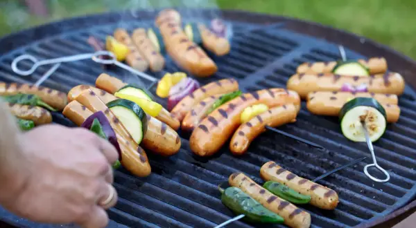 Grilla korv med grönsaker på en kolgrill. Röken stiger från de saftiga korvarna och grönsakerna som får fina grillränder.