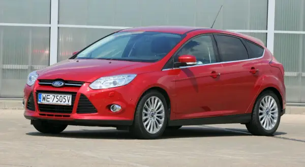 Czerwony Ford Focus z włączonymi światłami mijania, zaparkowany przed nowoczesnym budynkiem.