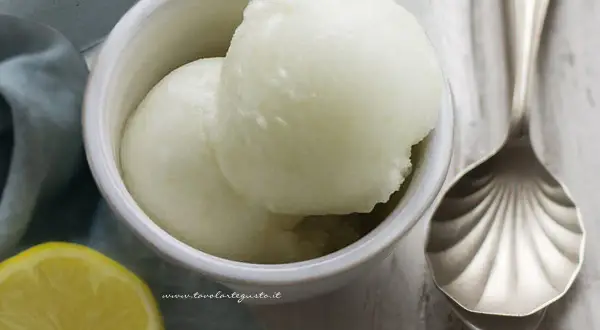 Due palline di sorbetto al limone senza albume in una ciotola bianca, con una fetta di limone e un cucchiaio decorato accanto.