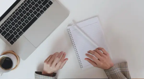 Ręce piszą notatki do CV po niemiecku przy laptopie, obok filiżanka kawy.