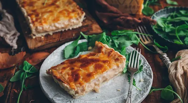 Una rebanada de tarta de cebolla dorada y cremosa, servida con rúcula fresca y un trozo de queso.
