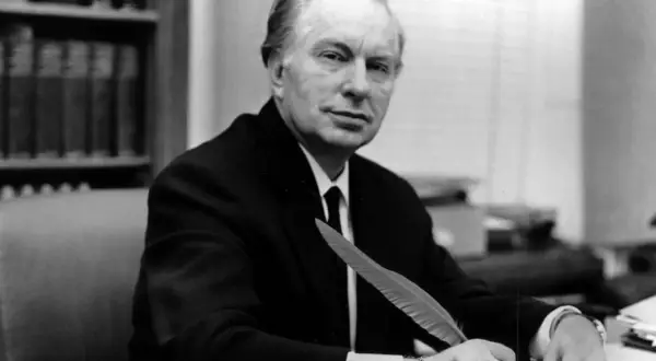 L. Ron Hubbard, twórca scjentologii, siedzi przy biurku z piórem w dłoni, gotów do pisania.