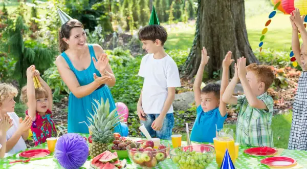 Radosne zabawy na festyn dla dzieci w ogrodzie. Dzieci w czapkach urodzinowych klaszczą, a na stole owoce i napoje.