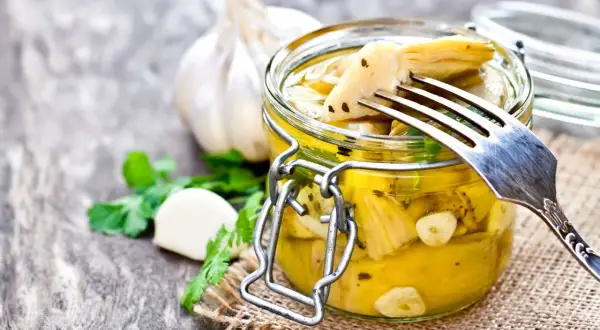 Deliziosi carciofini sott'olio fatti in casa, conservati in un barattolo di vetro con aglio e prezzemolo. Un forchettone ne solleva uno, pronto per essere gustato.
