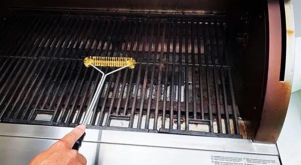 En hand rengör gallret på en gasolgrill under tak med en grillborste.