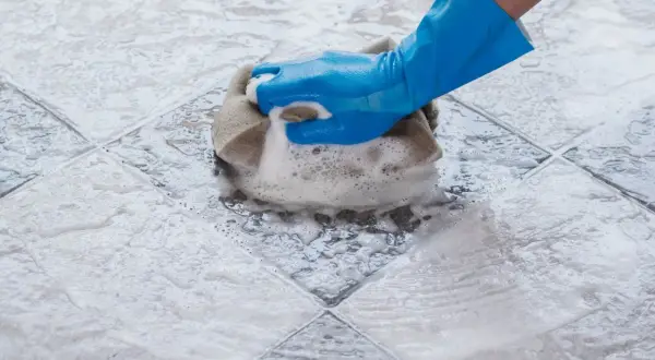 Main bleue frottant un carrelage avec une éponge, créant un voile de ciment savonneux.