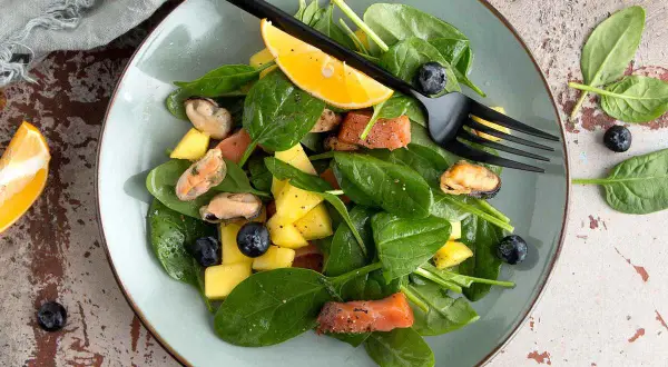 Insalata con mango, salmone, cozze e mirtilli, guarnita con spicchio d'arancia e forchetta nera.
