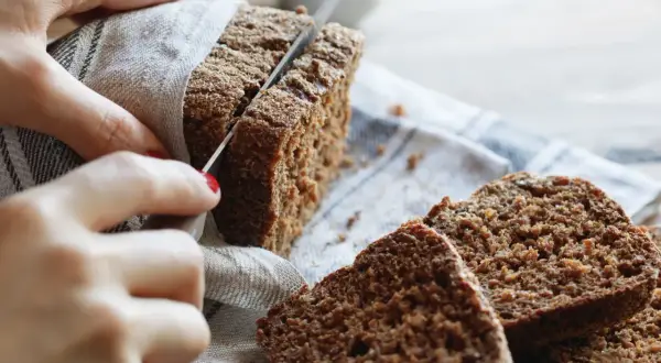 Hände schneiden ein dunkles, gesundes Roggenbrot. Mehrere Scheiben liegen auf einem Leinentuch.
