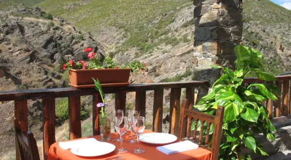 Mesa puesta para comer en Patones de Arriba, con vistas a un valle montañoso. Flores y plantas decoran el balcón.